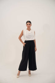 Woman wearing classic black wide-leg crepe pants with soft front pleats and elastic back, paired with a white sleeveless top and beige shoes, standing against a plain background.