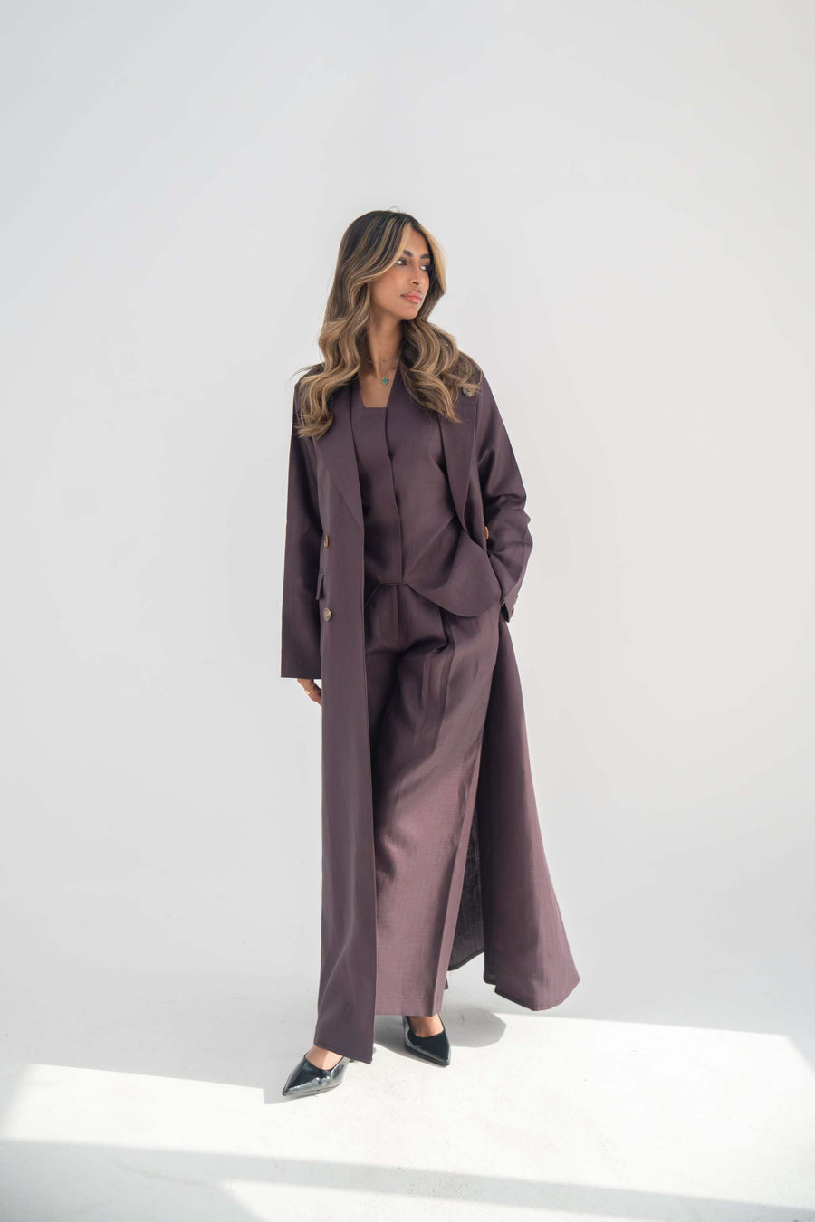 Woman wearing tailored blazer abaya in soft neutral tone with structured lapels, statement buttons, side flap pockets, and hidden inner pockets.