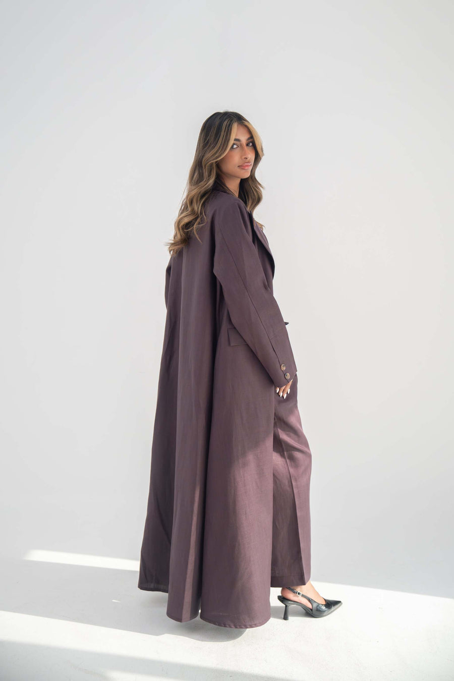 Woman wearing a tailored blazer abaya in soft neutral twill linen blend with structured lapels and statement buttons, standing against a plain background.
