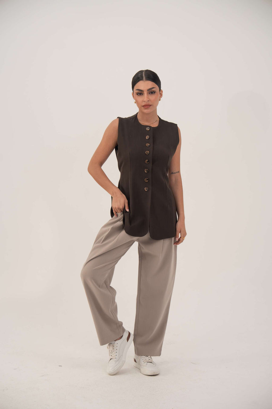 Woman wearing beige Truffle Crepe Pants with a black sleeveless buttoned top and white sneakers, posing against a plain background.