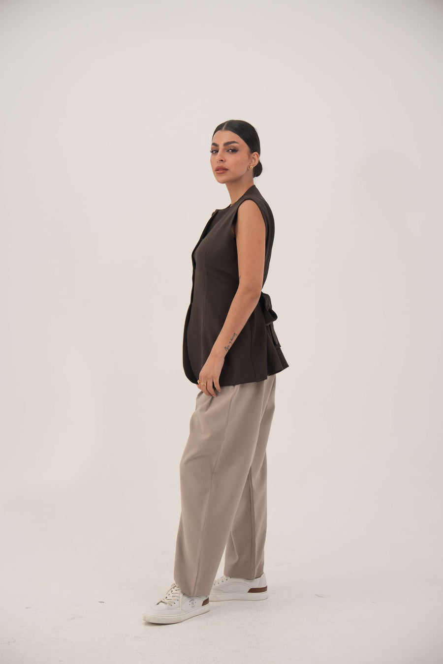 woman wearing beige truffle crepe pants with black sleeveless top and white sneakers standing against white background