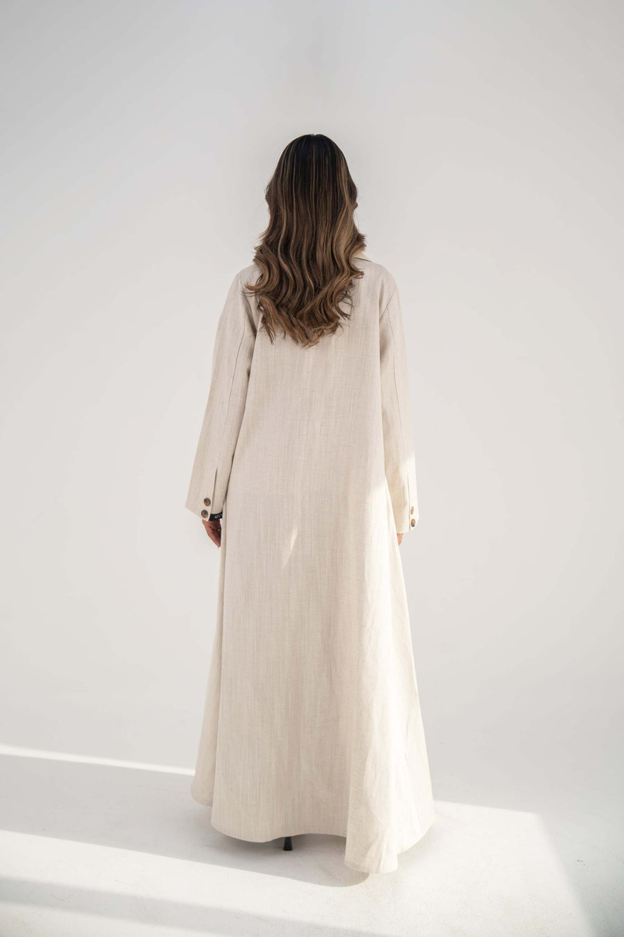 Back view of a woman wearing a warm beige blazer-style abaya made from soft twill linen blend with a flowy cut and long sleeves.