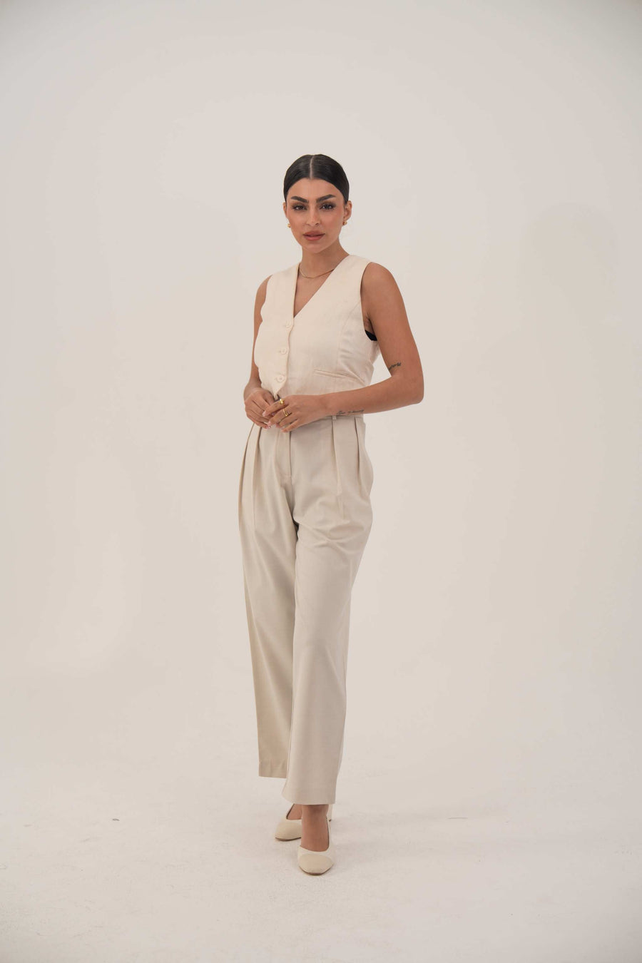 Woman wearing a sleeveless beige crepe vest with front buttons and matching beige pants in a minimalist setting