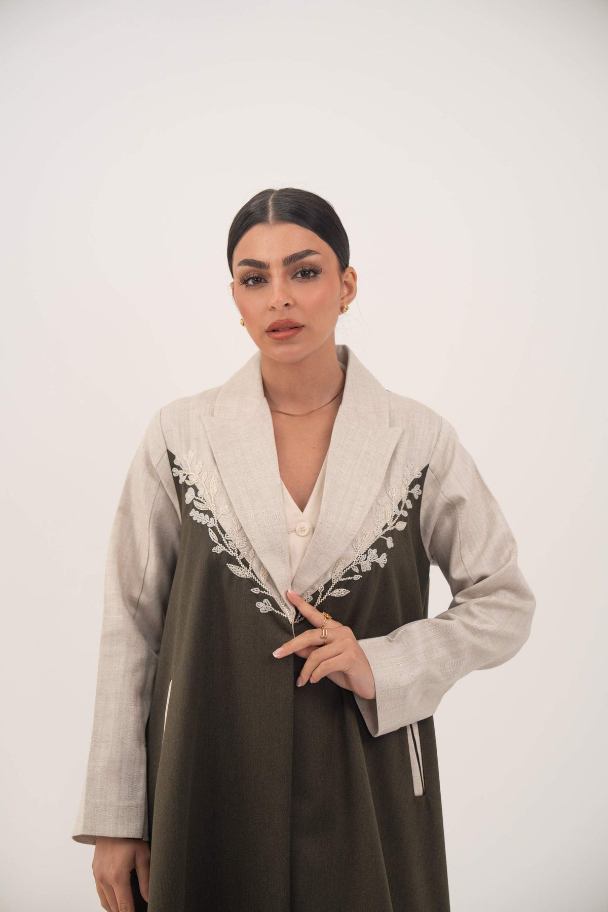 Olive green twill abaya with beige linen panels and white leaf embroidery on chest, featuring notched lapels, full-length cut, linen sleeve panels, shoulder pads, and beige-trimmed side slit pockets.
