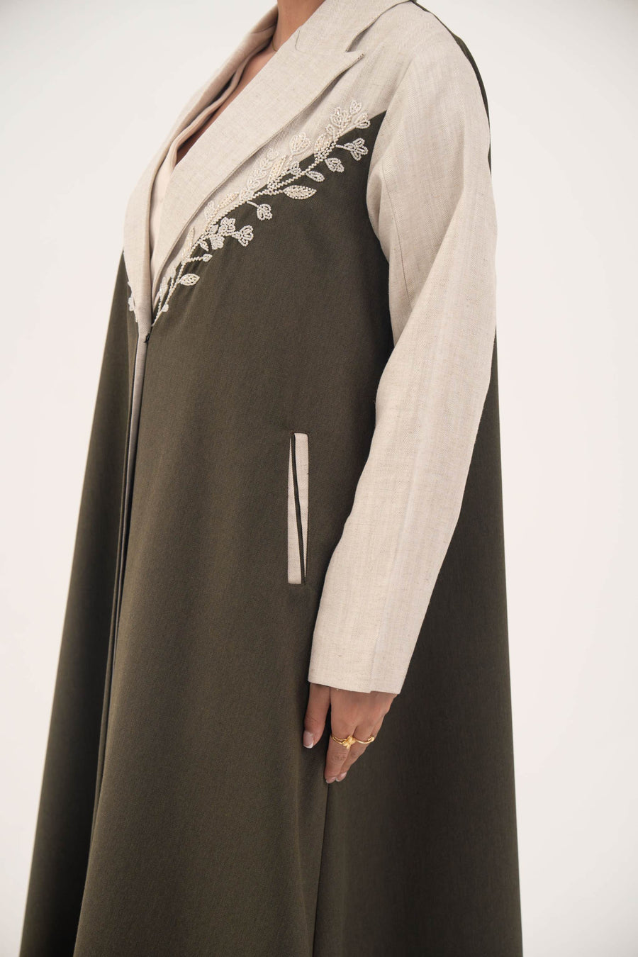 Deep olive green abaya with beige linen panels, white leaf embroidery, notched lapels, full-length cut, and beige-trimmed side slit pockets.