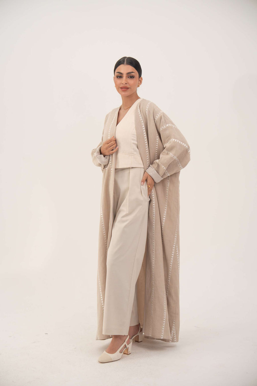Woman wearing a creamy beige airy linen abaya with tonal embroidery, soft chiffon cuffs, open front, and classic bisht silhouette.