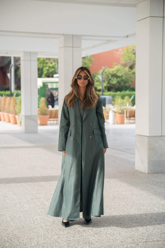 Woman wearing dark green linen blazer abaya with brown trim and buttons, standing outdoors.