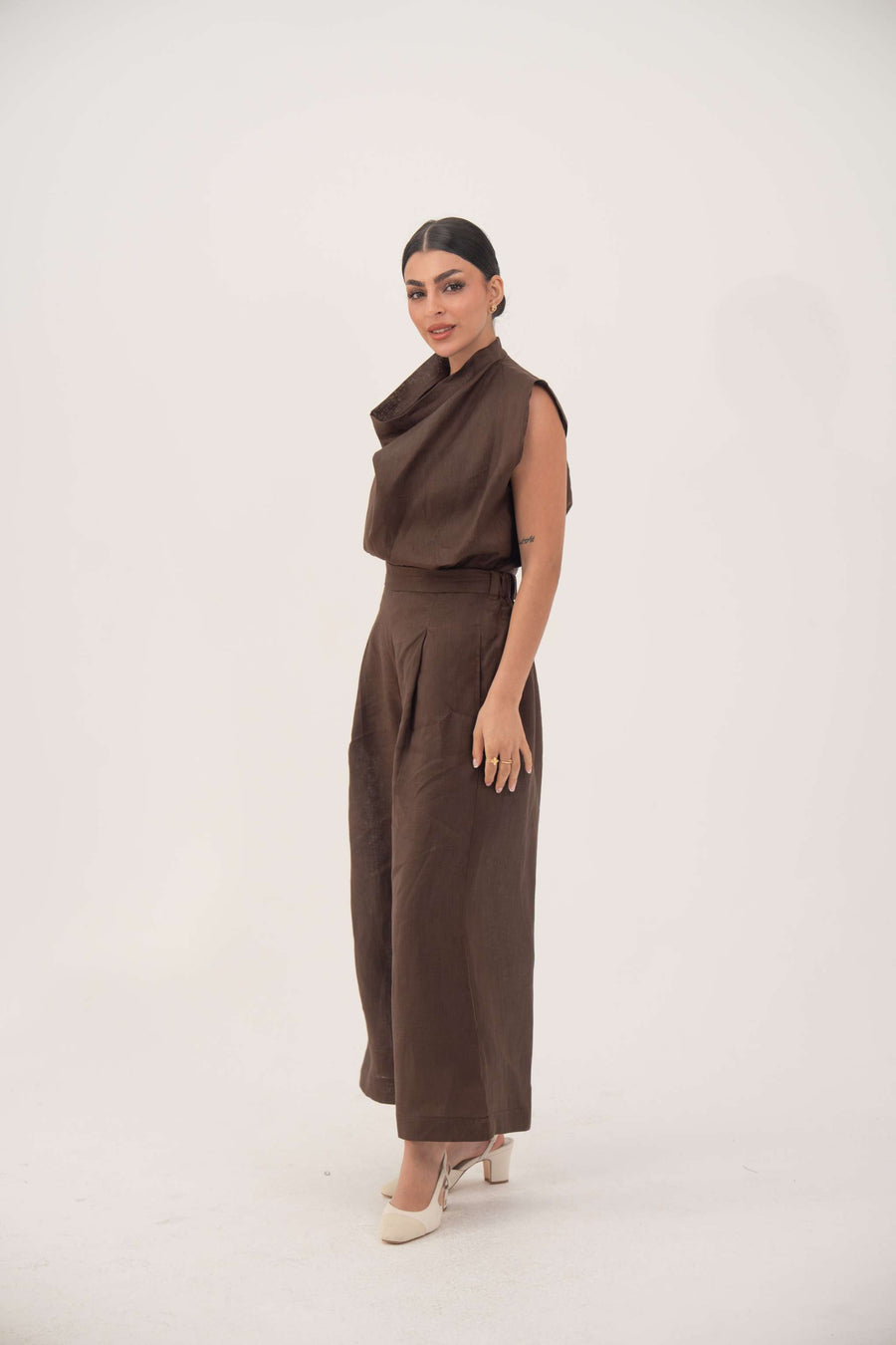 Woman wearing brown linen pleated sleeveless blouse and matching pants with draped neckline and elegant pleats.
