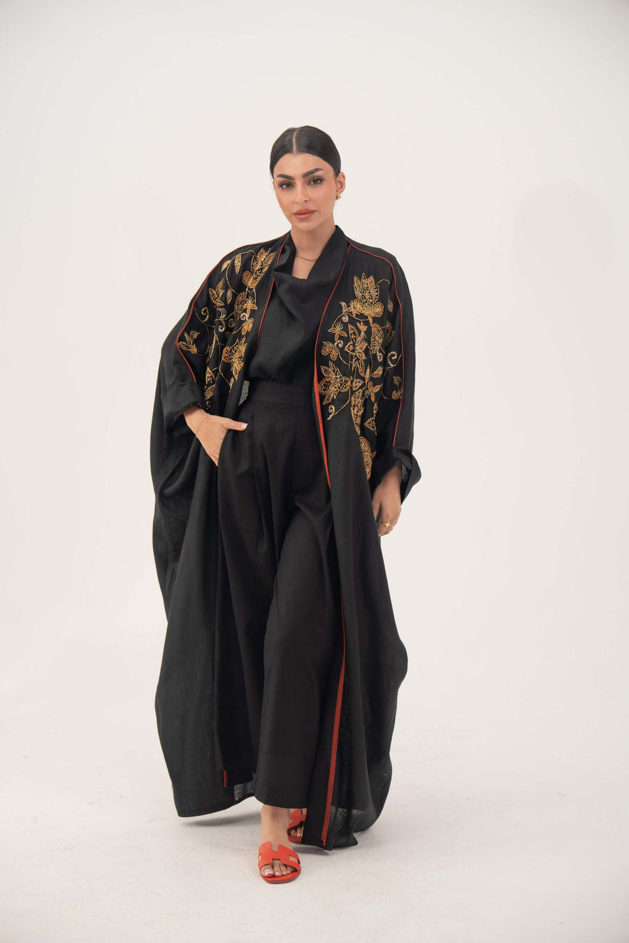 Woman wearing black Zureya bisht abaya with gold and bronze embroidery and orange trim, styled with wide sleeves and relaxed silhouette.