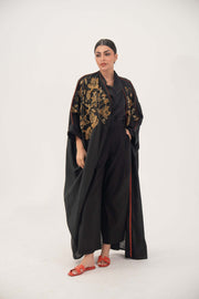 Black bisht abaya with golden bronze embroidery and orange trim, lightweight linen, loose open-front design, worn with black outfit and red sandals.
