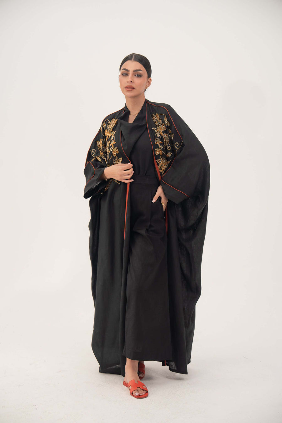 Woman wearing a black linen bisht abaya with gold and bronze embroidery and orange trim, featuring loose wide sleeves and a round neckline.
