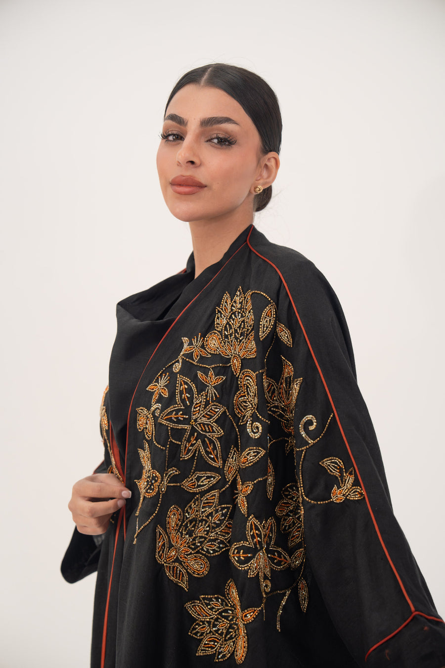 Black linen bisht abaya with golden and bronze floral embroidery and orange seam detailing, worn by a woman with a sleek hairstyle.