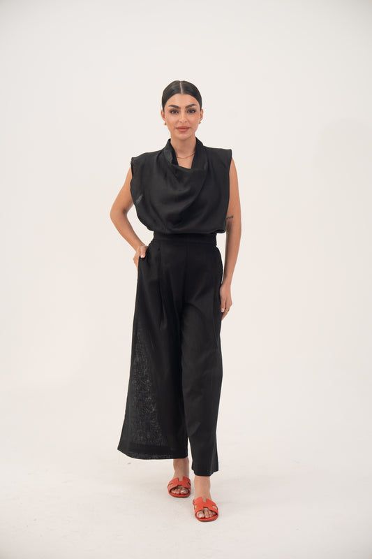 Woman wearing a sleeveless black linen blouse with draped neckline and pleats paired with black wide-leg pants and orange sandals.