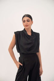Woman wearing a sleeveless black Noir linen blouse with a draped neckline and pleated detailing.