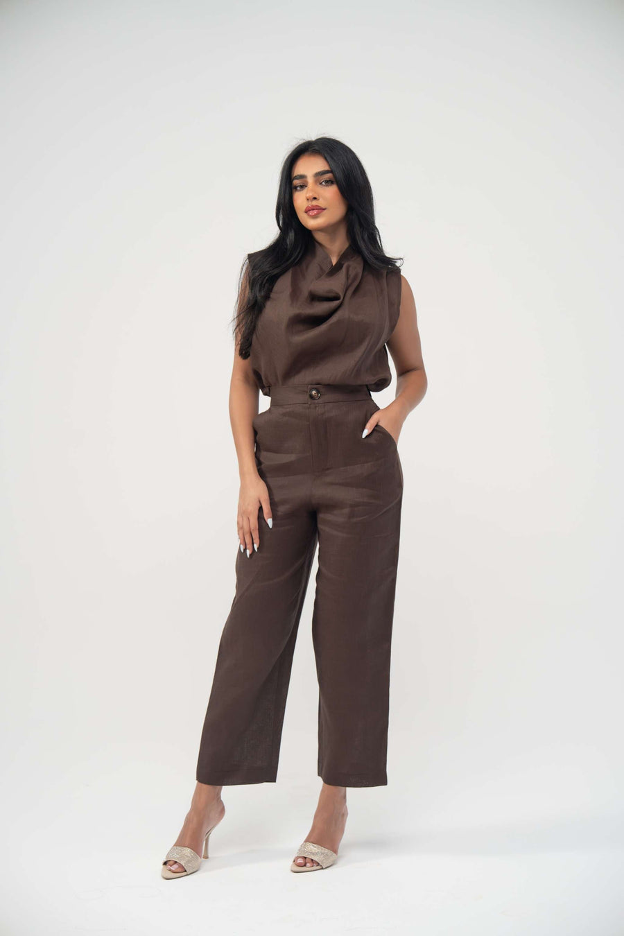 Woman wearing sleeveless warm brown espresso linen blouse with draped neckline and light pleats paired with matching pants and beige heels
