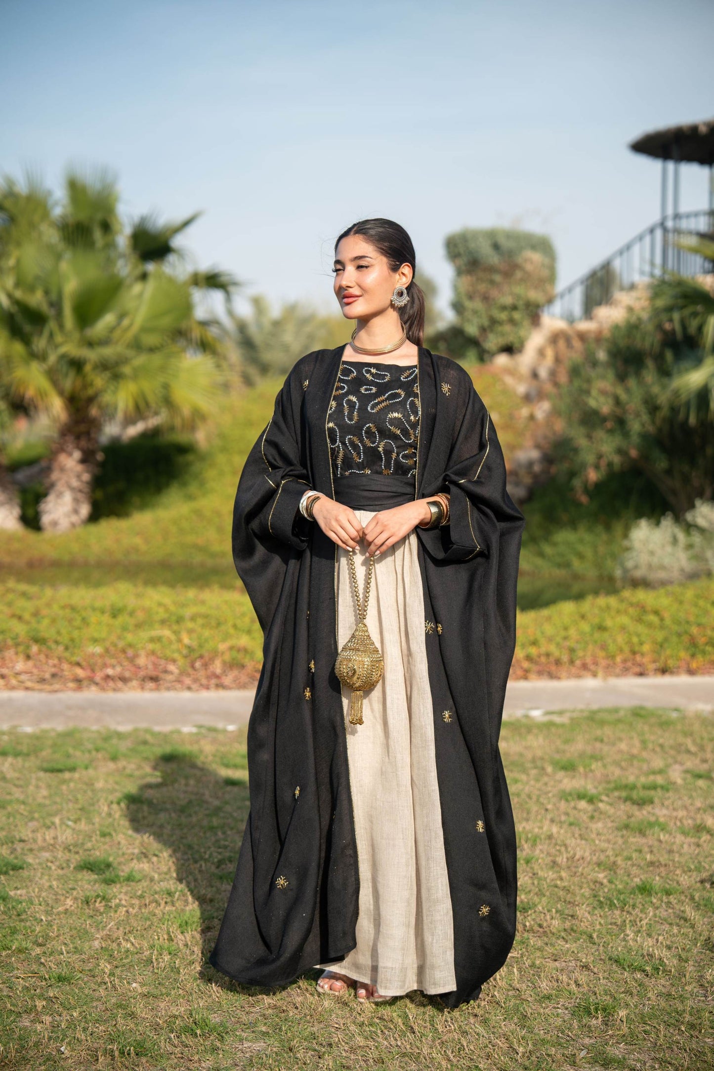 luxurious black bisht with gold embellishments over two-tone jumpsuit with black bodice and beige trousers on woman outdoors