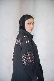 Side view of a woman wearing a full-length black linen abaya with multicolor traditional-inspired embroidery on the shoulders, sleeves, and back