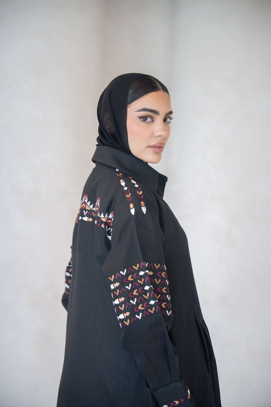 Side view of a woman wearing a full-length black linen abaya with multicolor traditional-inspired embroidery on the shoulders, sleeves, and back