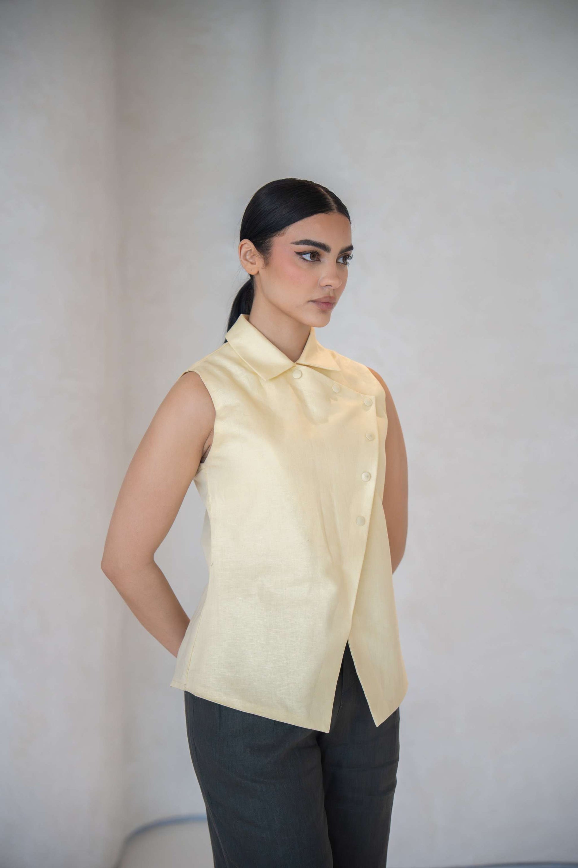Woman wearing a sleeveless butter yellow vest with an asymmetric front and fabric-covered buttons