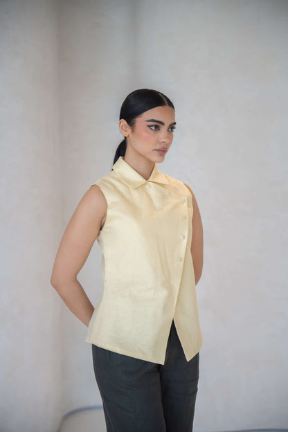 Woman wearing a sleeveless butter yellow vest with an asymmetric front and fabric-covered buttons