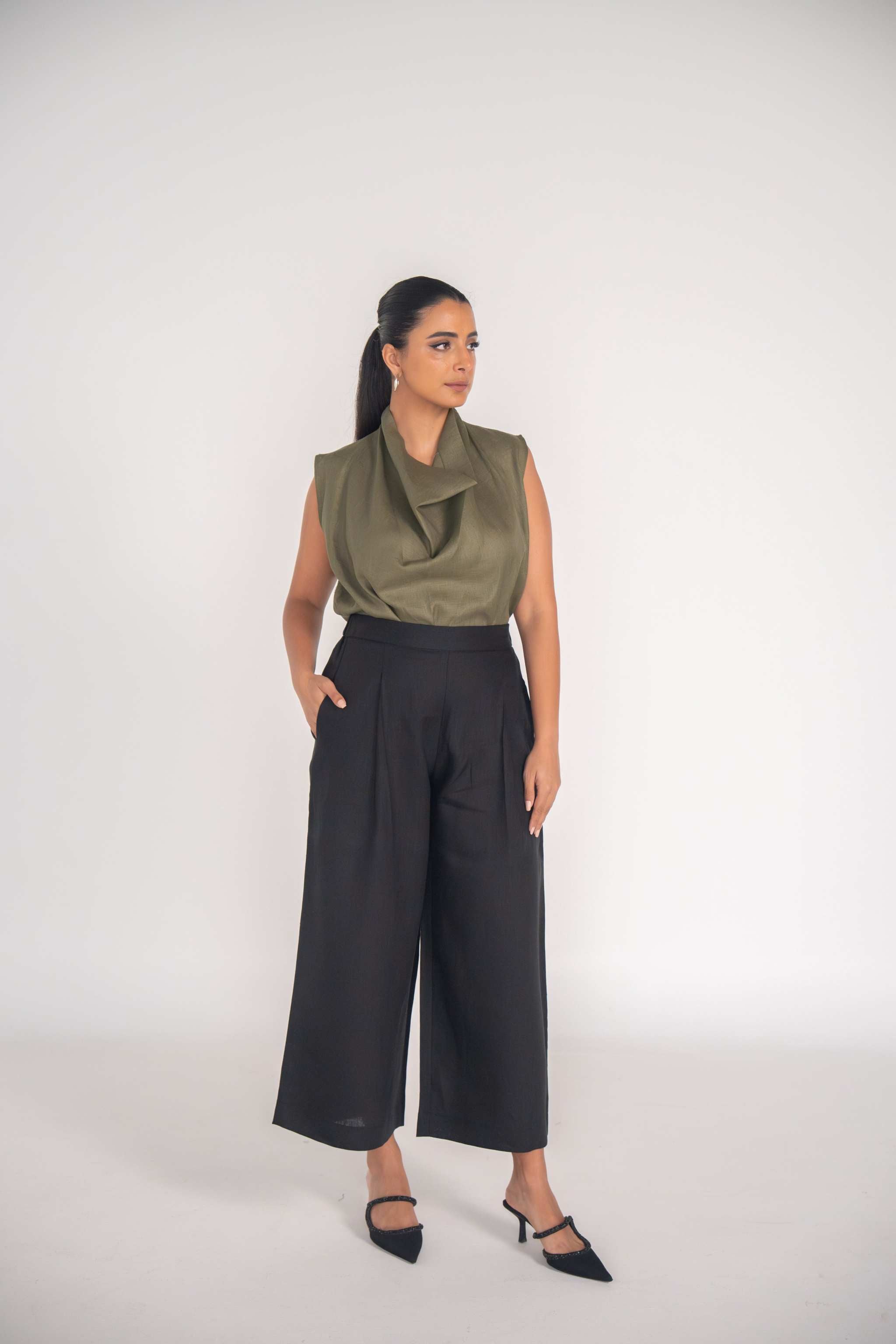 Woman wearing olive green sleeveless Raya blouse and black wide-leg trousers standing against plain background