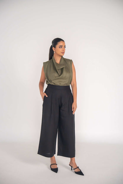 Woman wearing olive green sleeveless Raya blouse and black wide-leg trousers standing against plain background