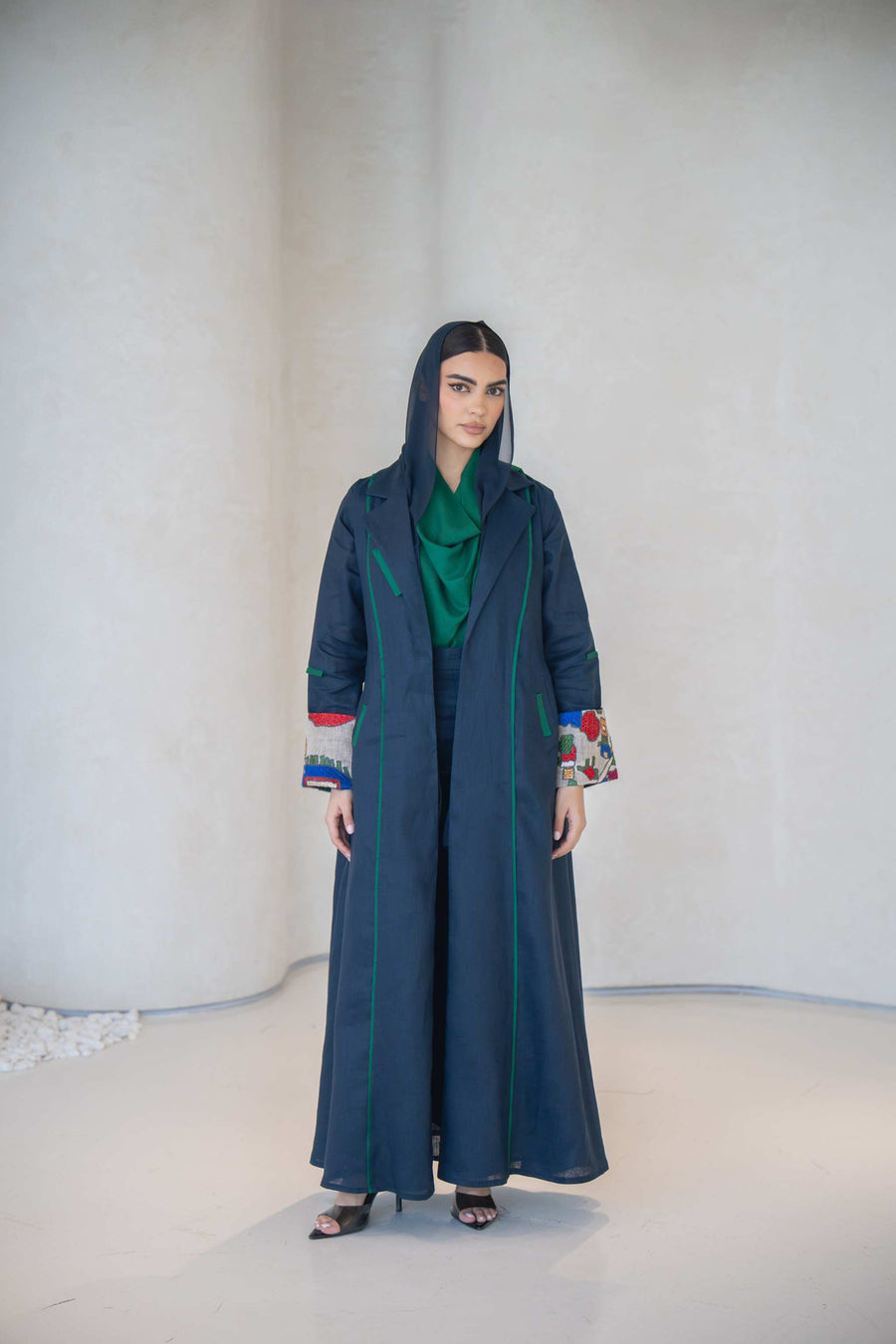 Woman wearing a deep navy linen abaya with emerald green piping and multicolor embroidered cuffs, featuring a modern open-front design and tailored silhouette.