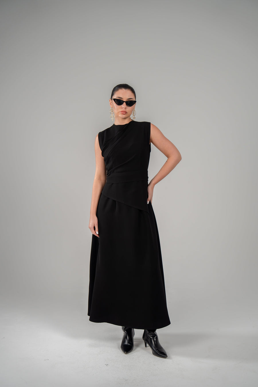 Woman wearing luxury black crepe dress with pleated neckline and matching belt against grey background