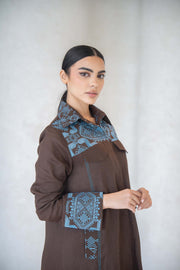 Woman wearing a calm brown abaya with blue vertical embroidery on collar and cuffs, featuring a faux flap pocket.