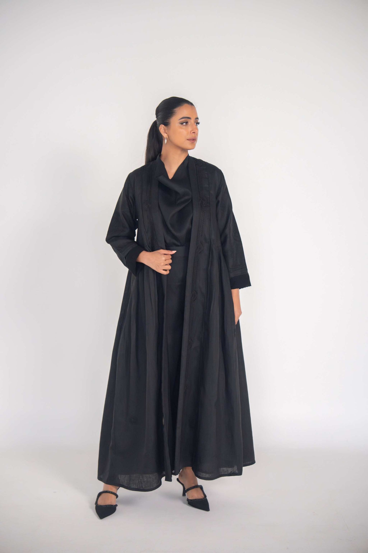 Woman wearing a black linen abaya with pleated sides and subtle black-on-black embroidery, styled with black shoes.