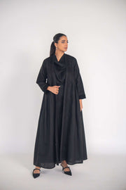 Woman wearing a black linen abaya with pleated sides and subtle black-on-black embroidery, styled with black shoes.
