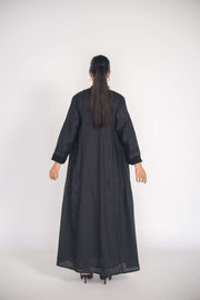 Back view of a woman wearing a refined black linen abaya with soft pleats and subtle black-on-black embroidery, featuring full lining and a flowy silhouette.