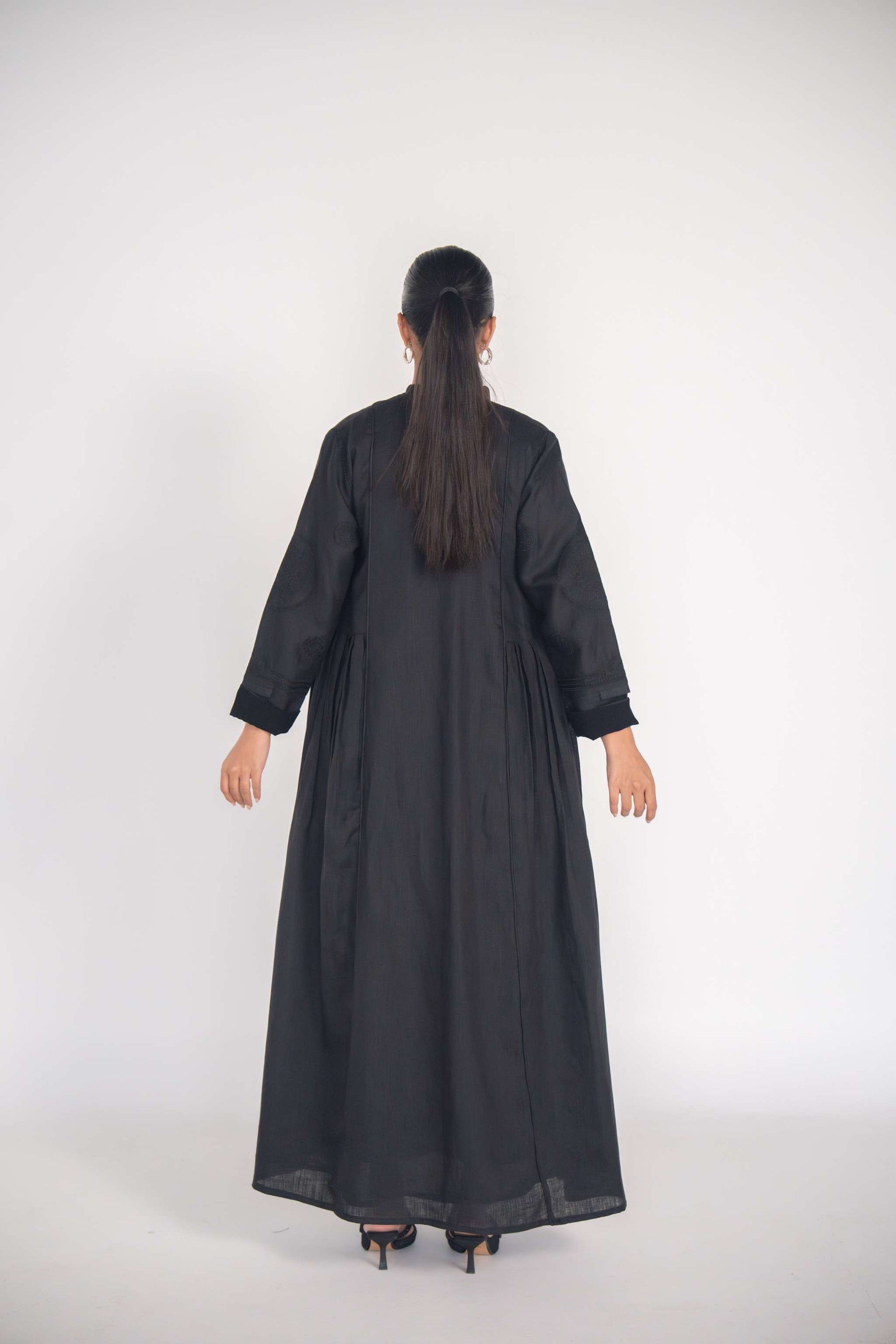 Back view of a woman wearing a refined black linen abaya with soft pleats and subtle black-on-black embroidery, featuring full lining and a flowy silhouette.