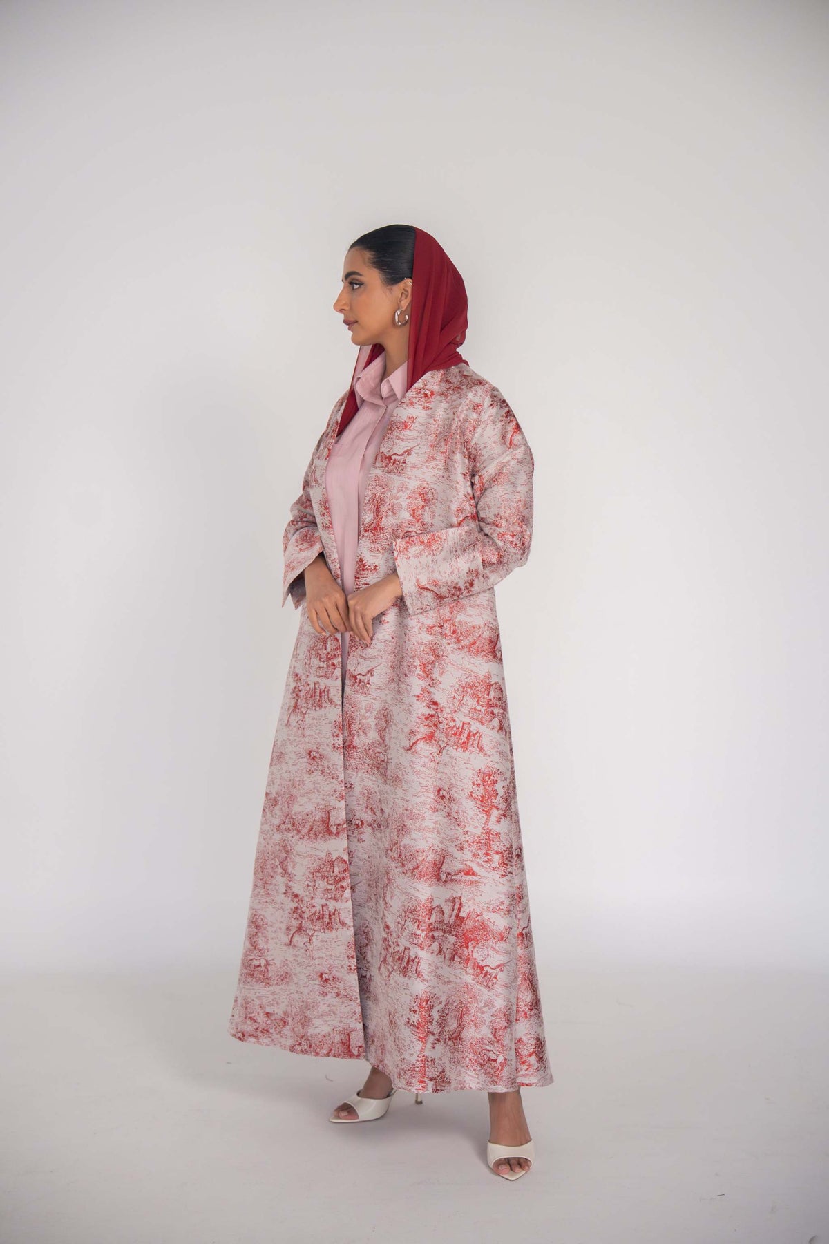 Woman wearing Cherry Haneen jacquard abaya with red Toile de Jouy countryside pattern on off-white background, styled with matching headscarf and heels.