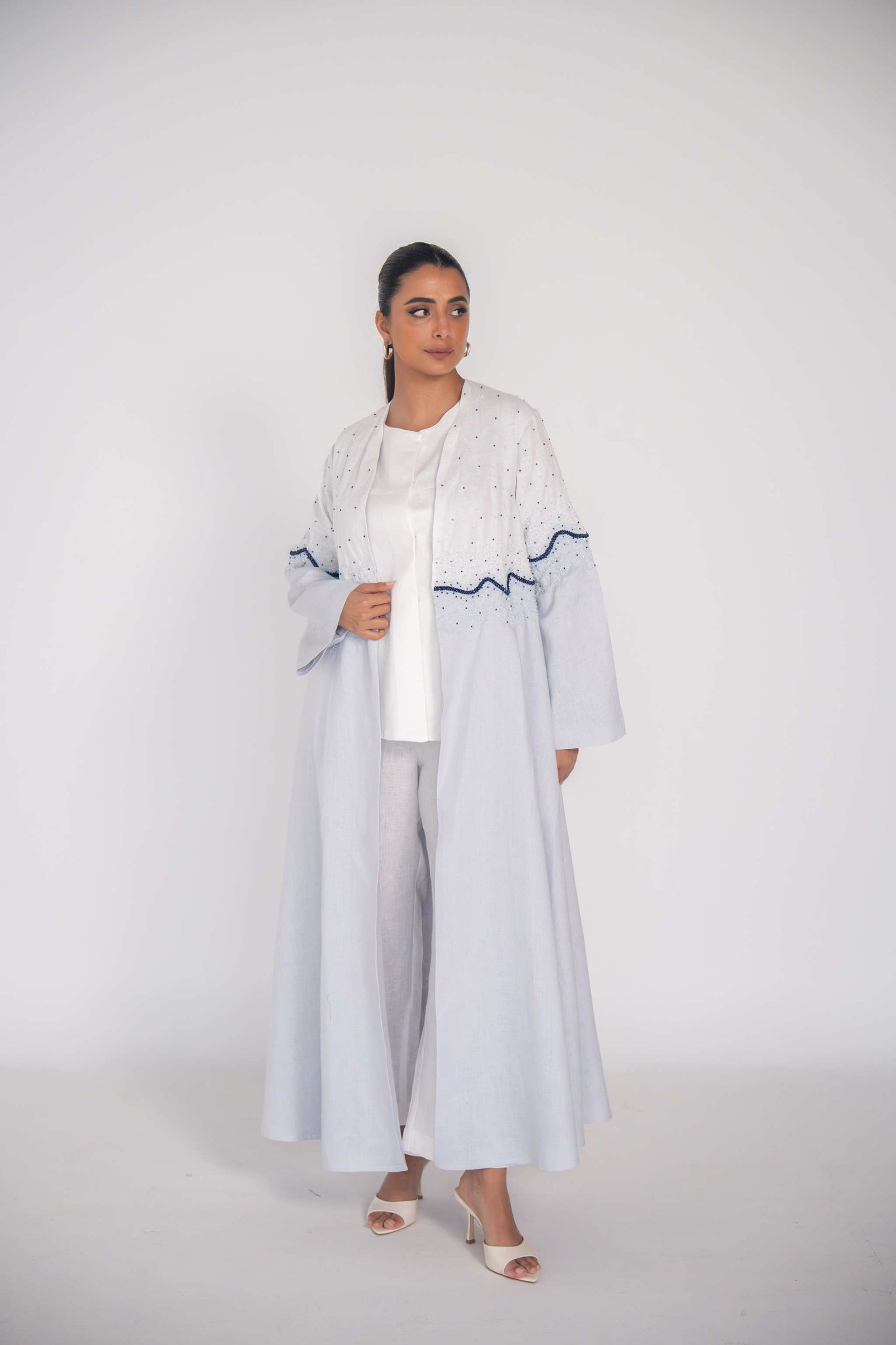 woman wearing a soft blue refined linen abaya with hand-applied white and navy beadwork in a wave pattern across the bodice and sleeves
