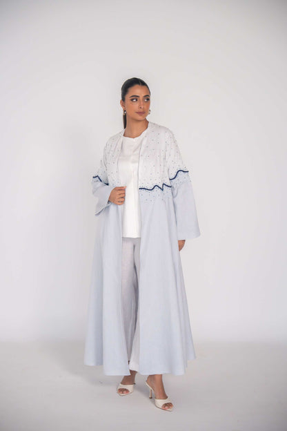 woman wearing a soft blue refined linen abaya with hand-applied white and navy beadwork in a wave pattern across the bodice and sleeves