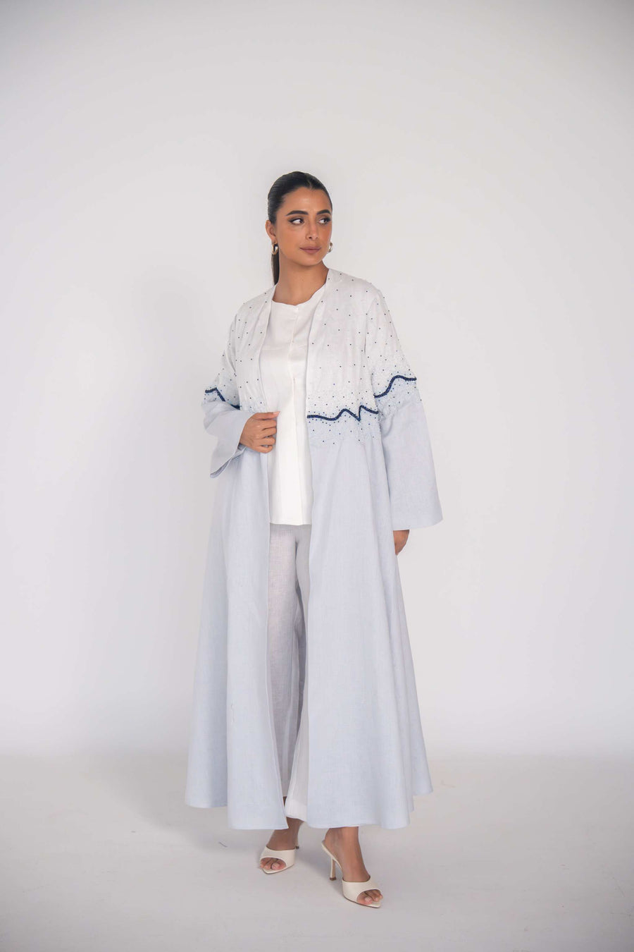 woman wearing a soft blue refined linen abaya with hand-applied white and navy beadwork in a wave pattern across the bodice and sleeves