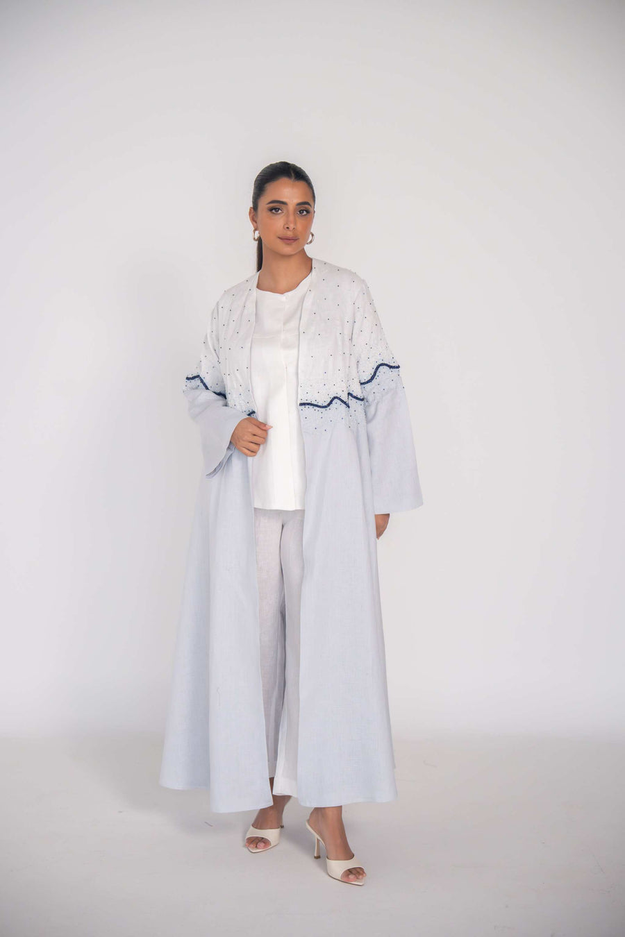 Woman wearing a light linen abaya with hand-applied blue and white beadwork in a wave pattern on bodice and sleeves, featuring a flowing cut and chiffon lining.