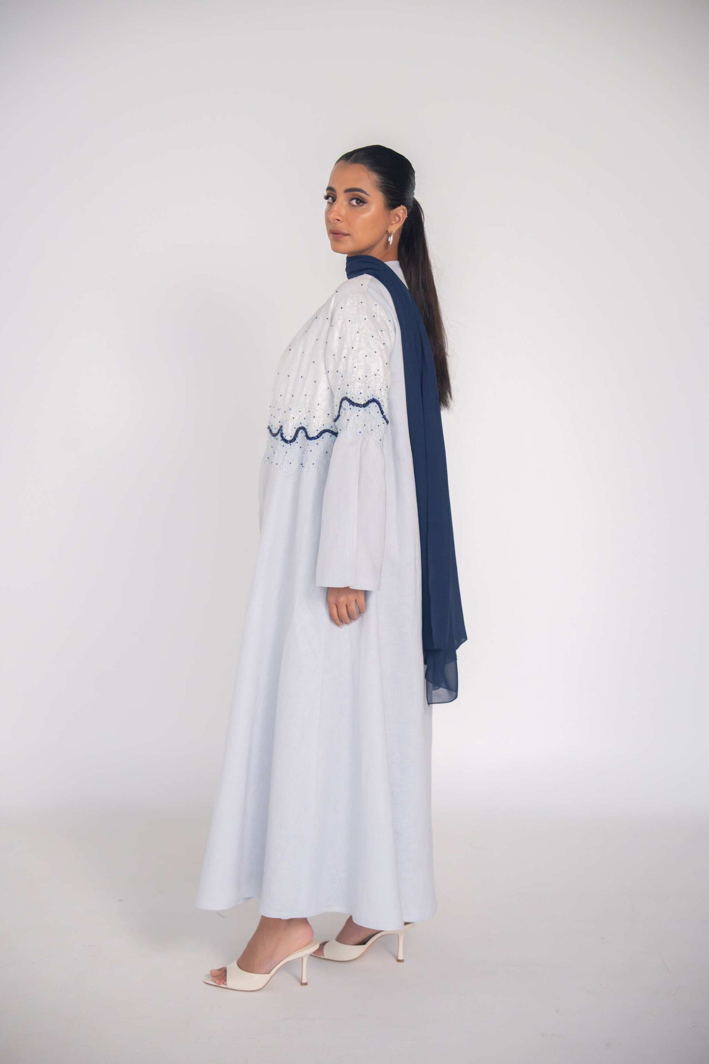 Woman wearing a white linen abaya with navy wave beadwork and chiffon lining, styled for warm weather.