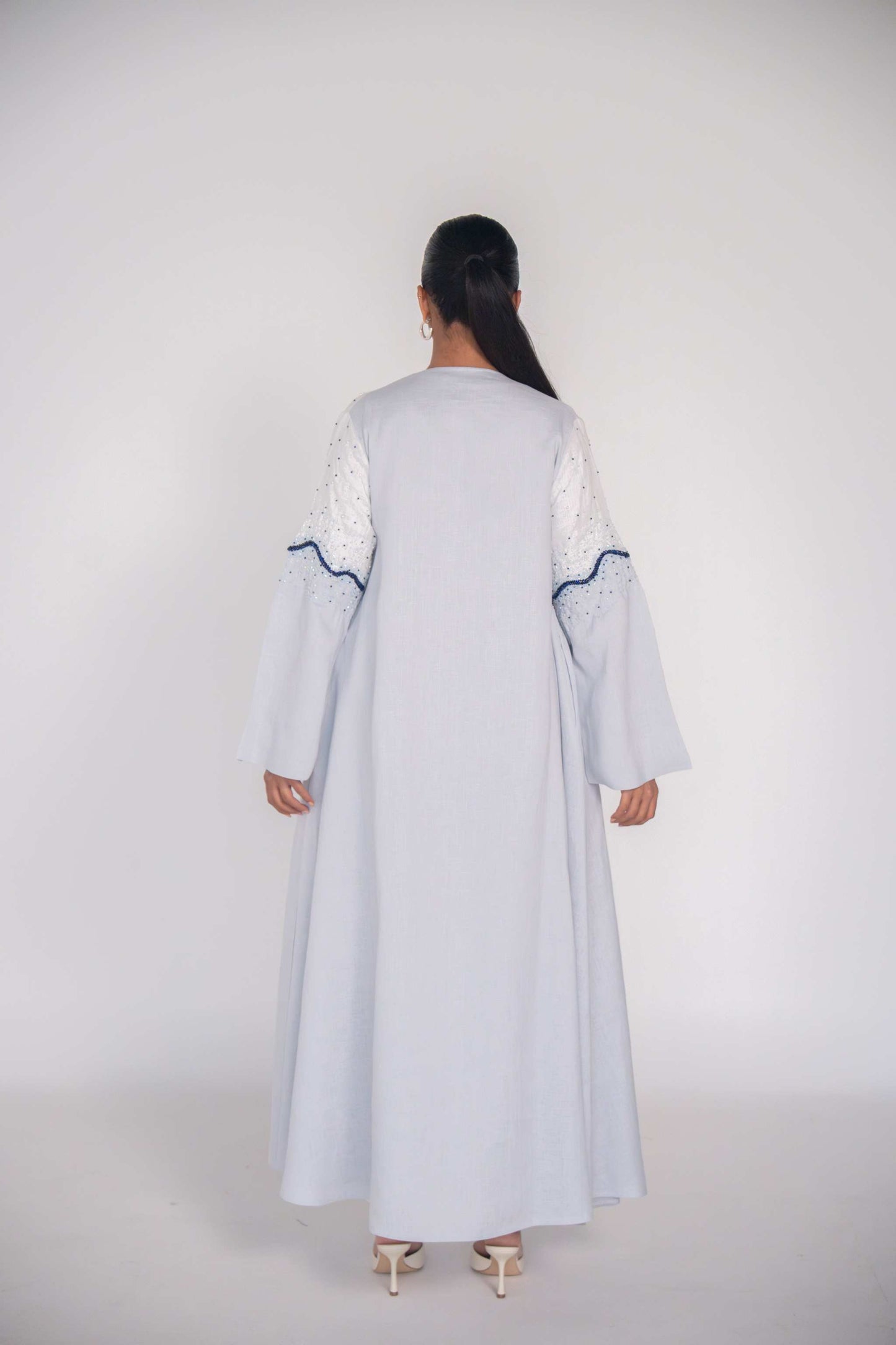 Back view of a light linen abaya with hand-applied white and navy beadwork on sleeves and bodice, featuring a flowing cut and chiffon lining for comfort.
