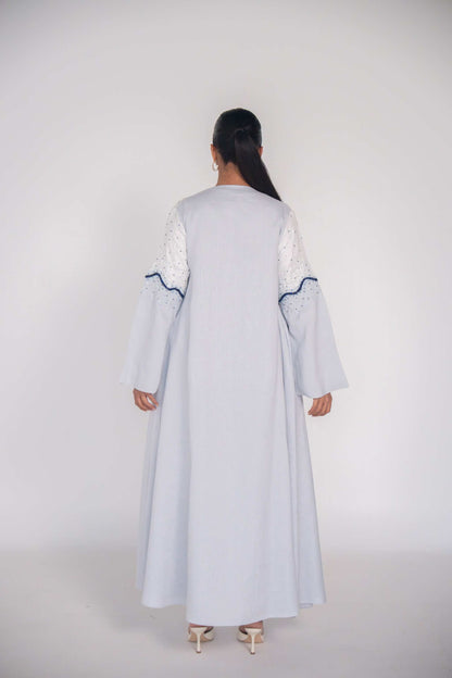 Back view of a light linen abaya with hand-applied white and navy beadwork on sleeves and bodice, featuring a flowing cut and chiffon lining for comfort.