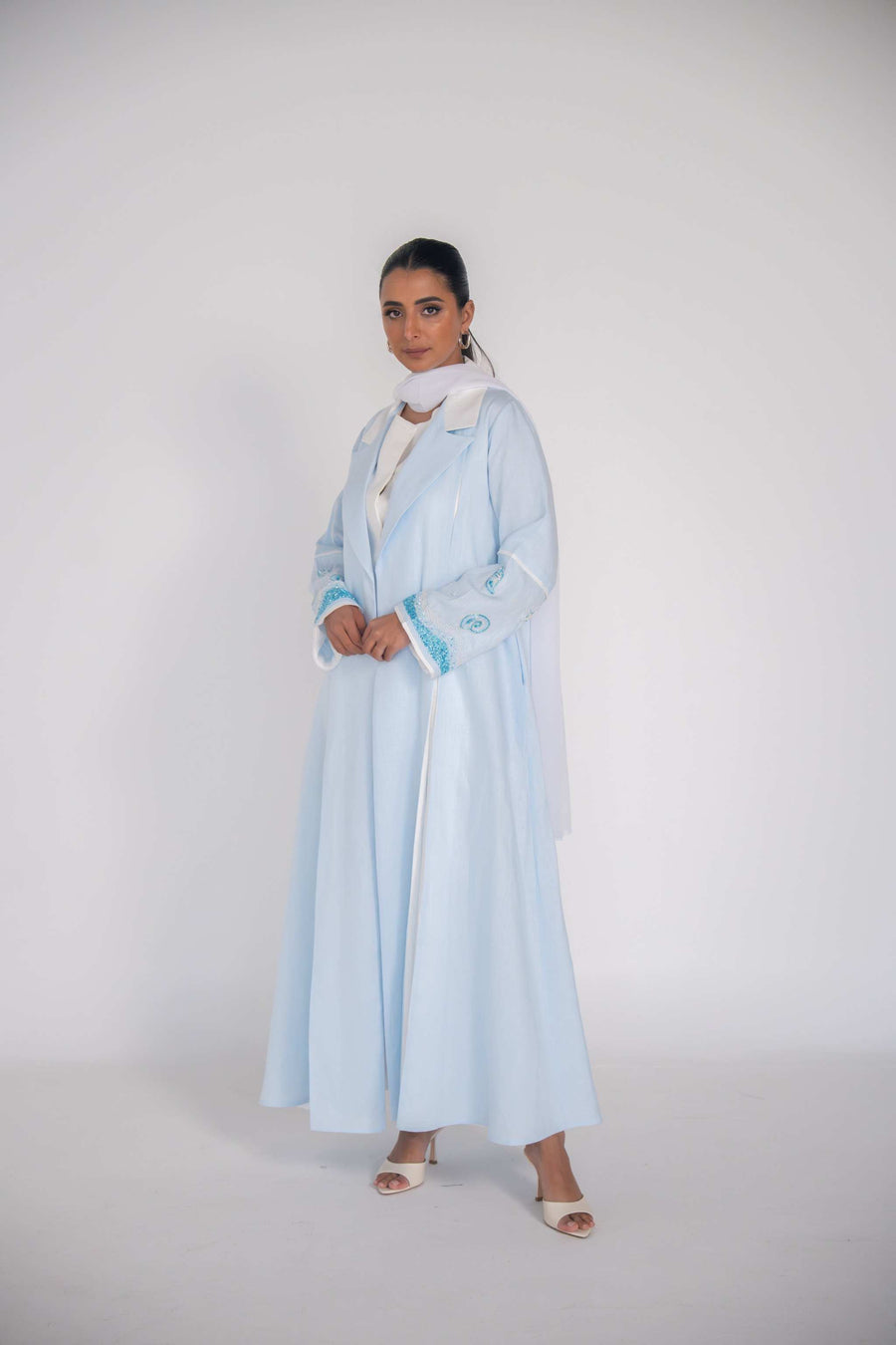 Woman wearing a baby blue abaya with tailored blazer collar and embroidered ocean-themed sleeves, paired with white scarf and heels.