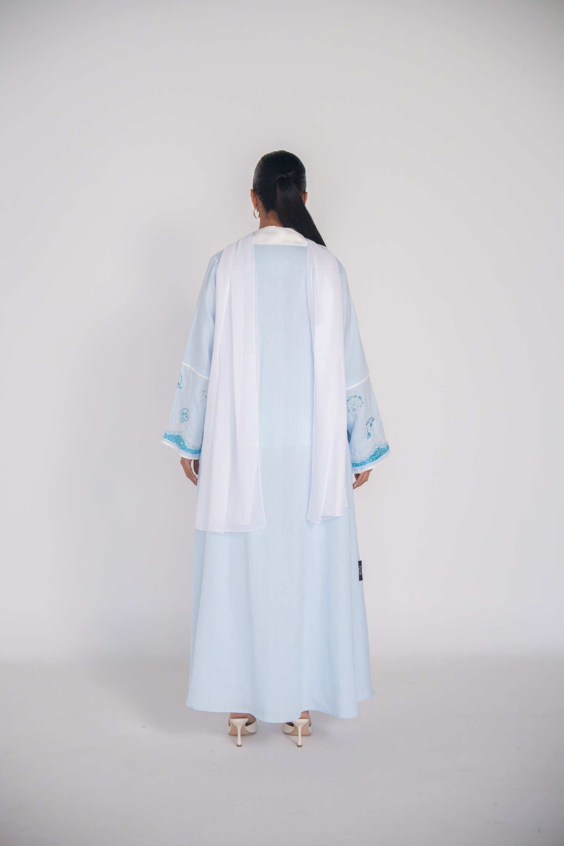 Woman wearing a baby blue abaya with tailored blazer collar and embroidered ocean-themed sleeves, paired with white scarf and heels.