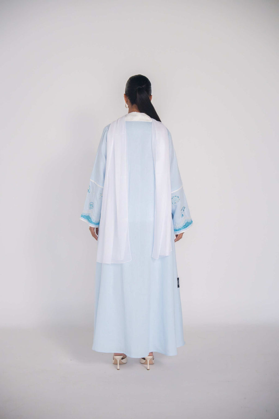 Woman wearing a baby blue abaya with tailored blazer collar and embroidered ocean-themed sleeves, paired with white scarf and heels.