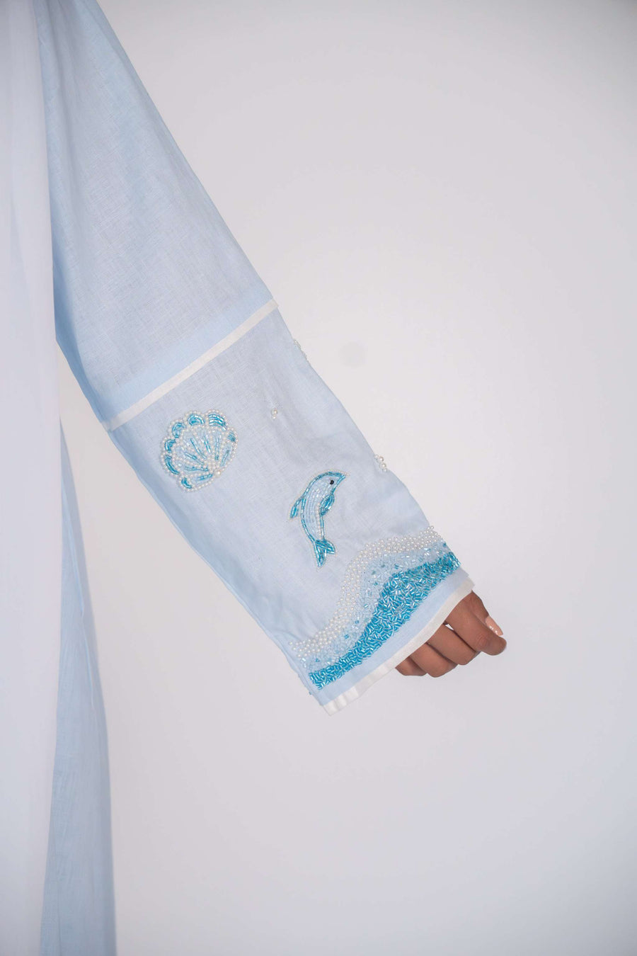 Woman wearing a baby blue abaya with tailored blazer collar and embroidered ocean-themed sleeves, paired with white scarf and heels.