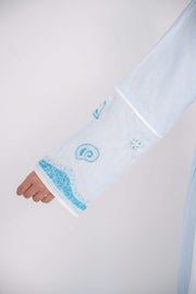 Woman wearing a baby blue abaya with tailored blazer collar and embroidered ocean-themed sleeves, paired with white scarf and heels.