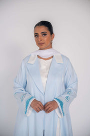 Woman wearing a baby blue abaya with tailored blazer collar and embroidered ocean-themed sleeves, paired with white scarf and heels.
