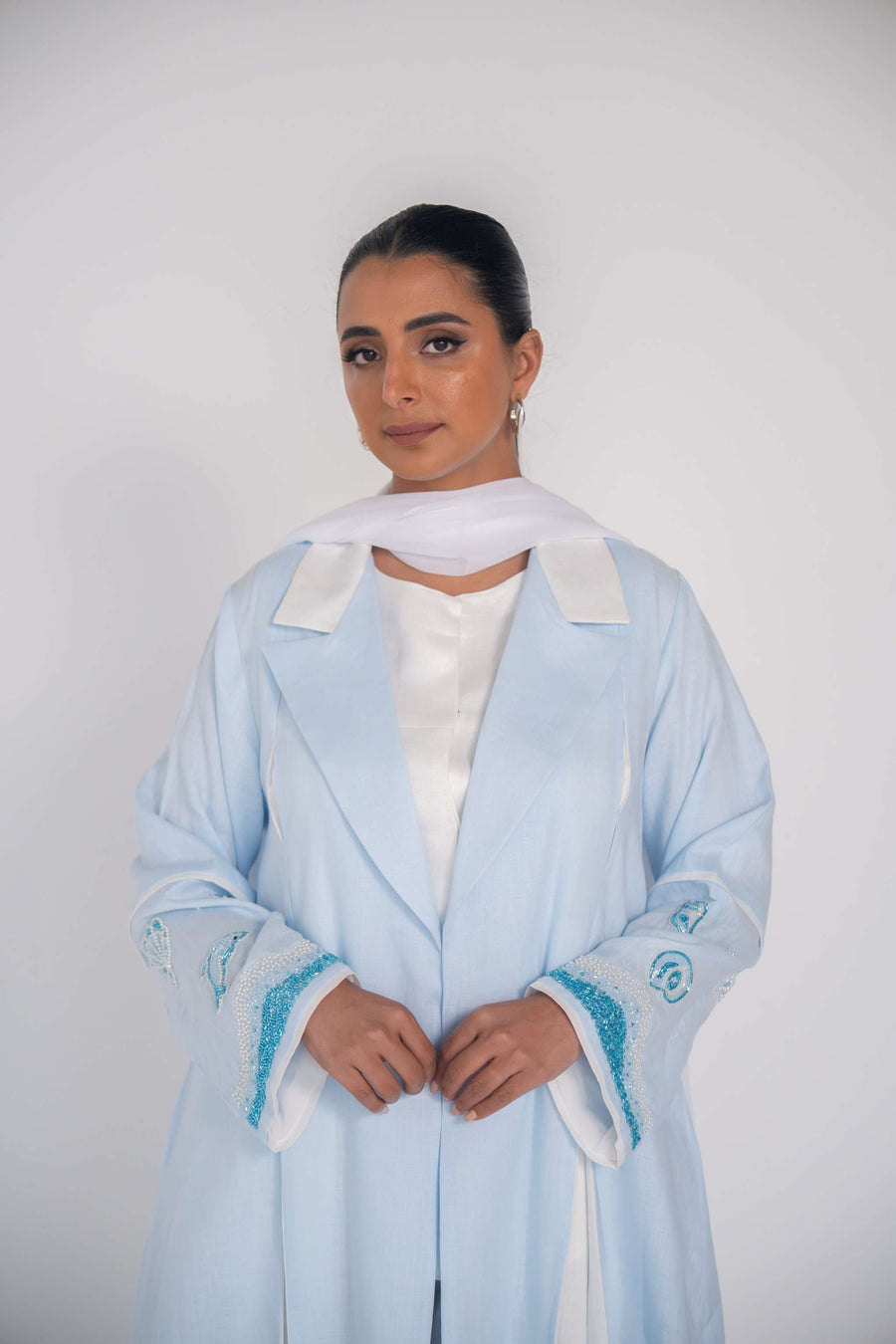 Woman wearing a baby blue abaya with tailored blazer collar and embroidered ocean-themed sleeves, paired with white scarf and heels.