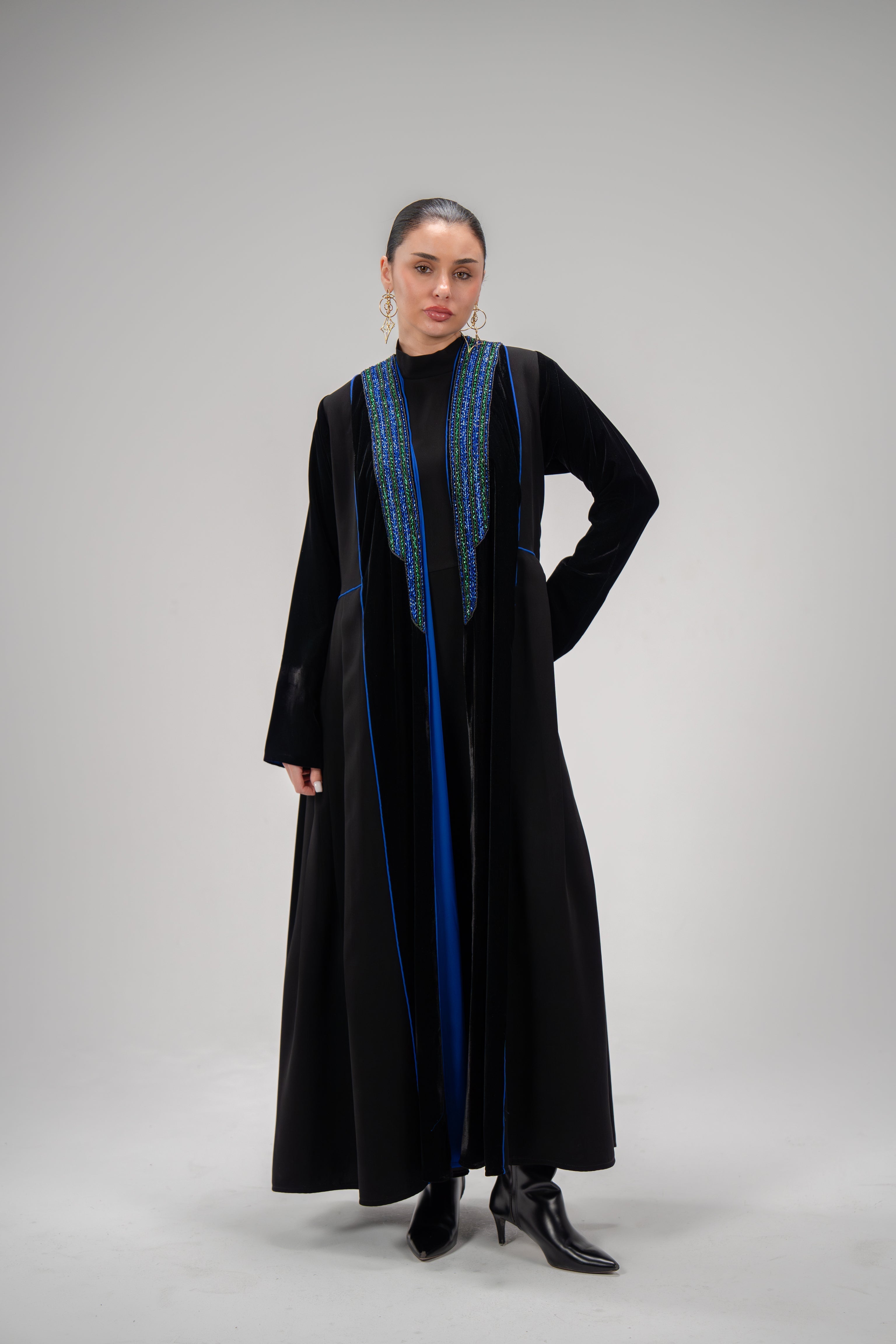 Sapphire abaya in black crepe and velvet with blue hand embroidery and side pleats, worn by a woman with black boots.
