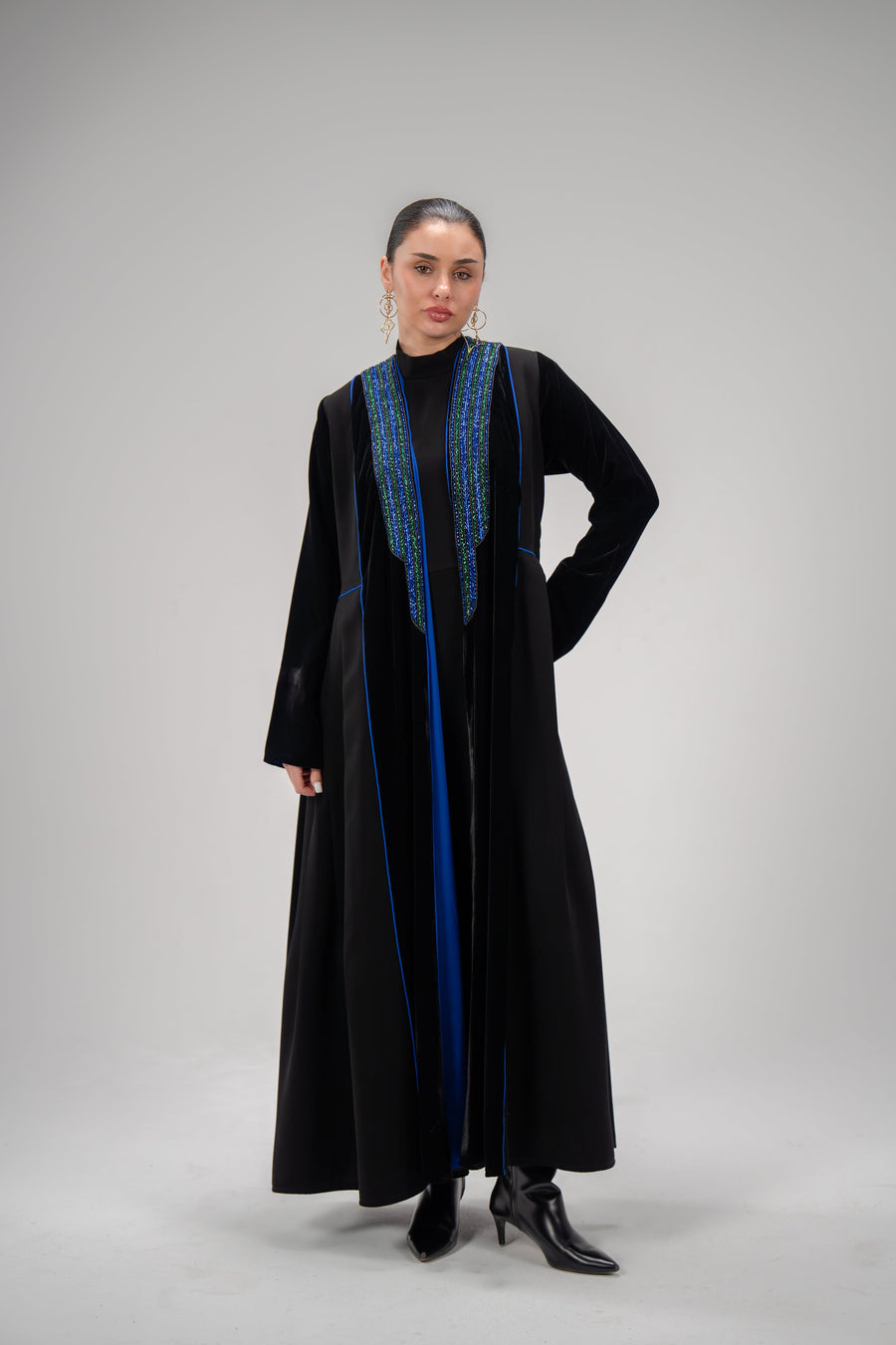 Sapphire abaya in black crepe and velvet with blue hand embroidery and side pleats, worn by a woman with black boots.